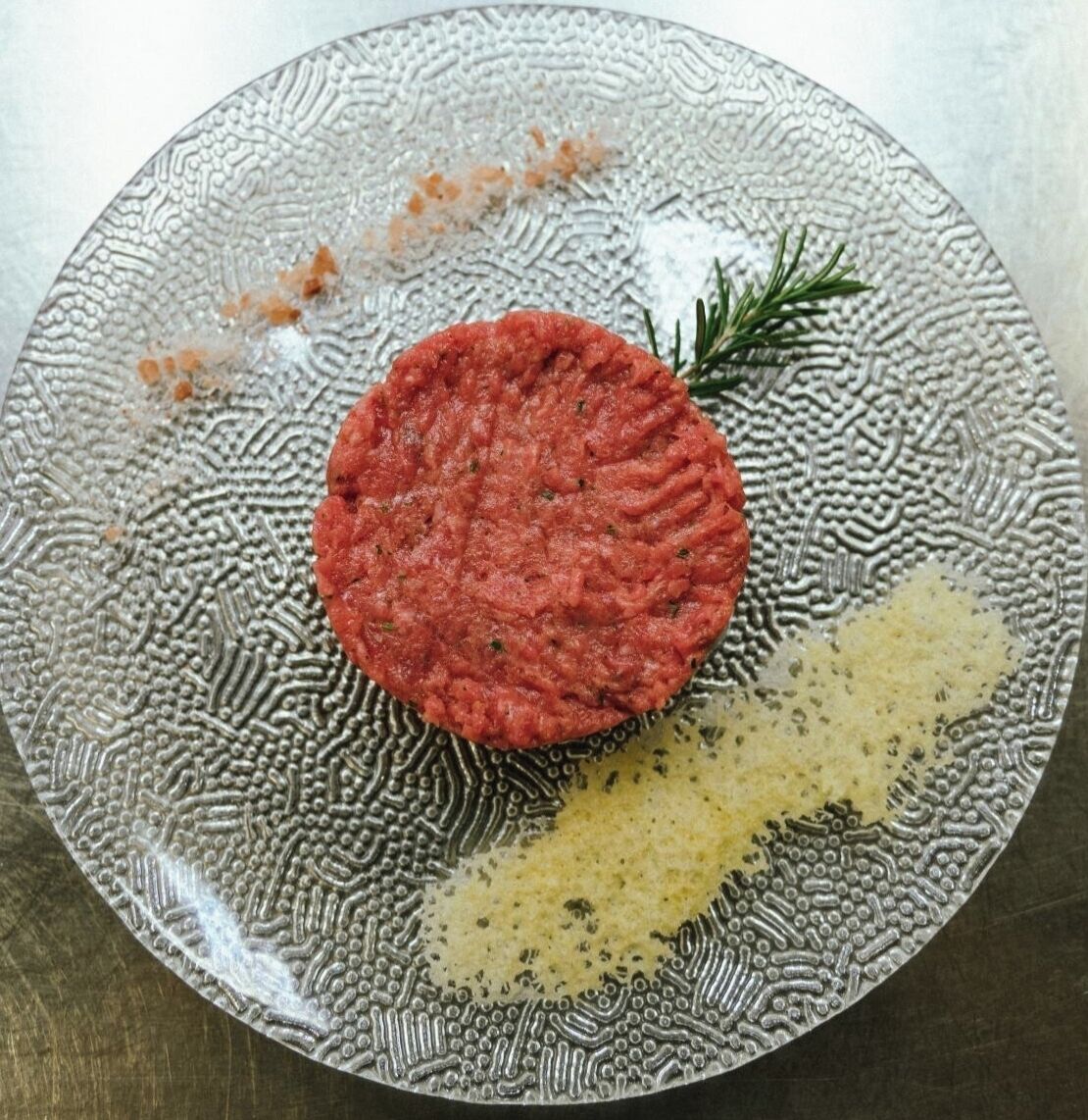 Une assiette ronde, à la texture particulière, sur laquelle on trouve une galette de viande au centre, une branche de romarin et une touche de sauce jaune.