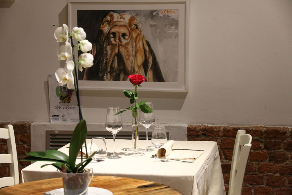 Mesa de jantar posta com toalha branca, copos de vinho, um vaso com orquídeas brancas e um quadro na parede.