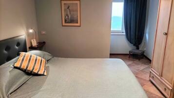 Een slaapkamer met een bed, een nachtkastje, een lamp, een ingelijst beeld aan de muur, en uitzicht buiten door een raam.