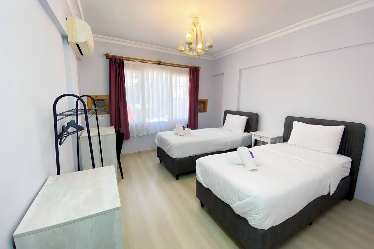 A hotel room with two beds, a desk, a chair, a radiator, and a window with curtains.