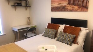 Una cama bien hecha con almohadillas estampadas, una mesita de noche con un florero, y un estante en la pared con libros y un objeto decorativo.