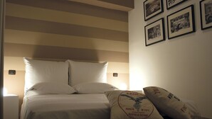 Een keurig opgemaakt bed met witte lakens en kussens, een nachtkastje met een lamp, en een muur met ingelijste foto's.