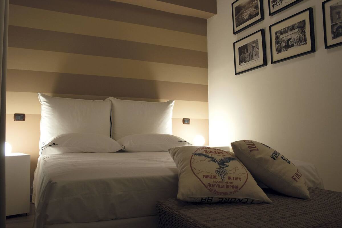 Een keurig opgemaakt bed met witte lakens en kussens, een nachtkastje met een lamp, en een muur met ingelijste foto's.