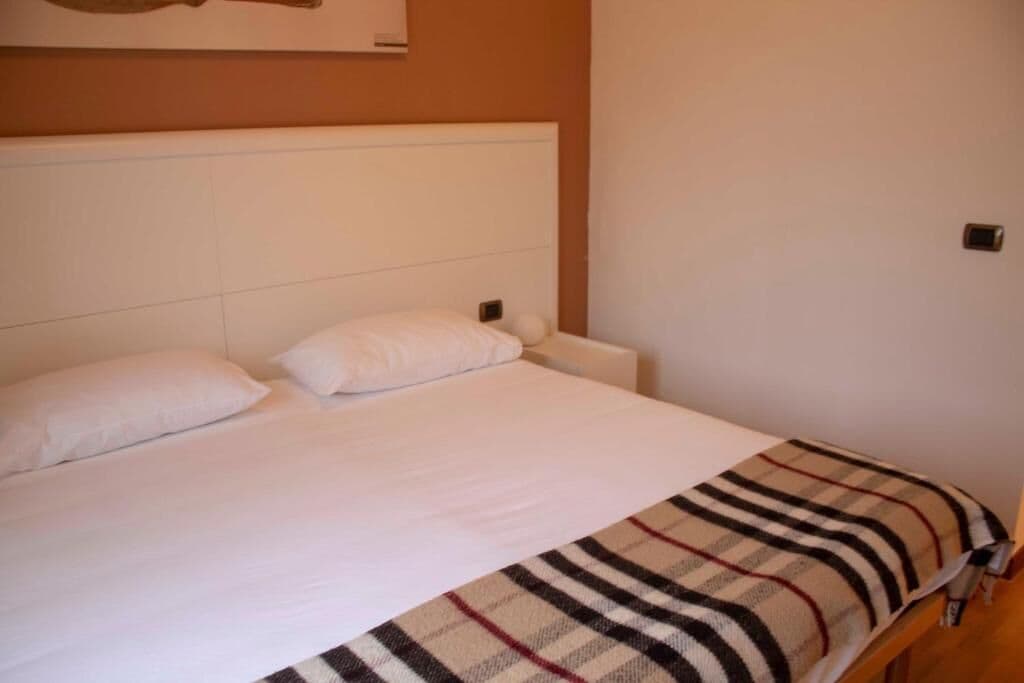 Een eenpersoonsbed met witte beddengoed en een geruit plaid, een wit hoofdbord en een effen muur met een kleine lichtschakelaar.