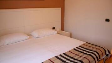 Een eenpersoonsbed met witte beddengoed en een geruit plaid, een wit hoofdbord en een effen muur met een kleine lichtschakelaar.