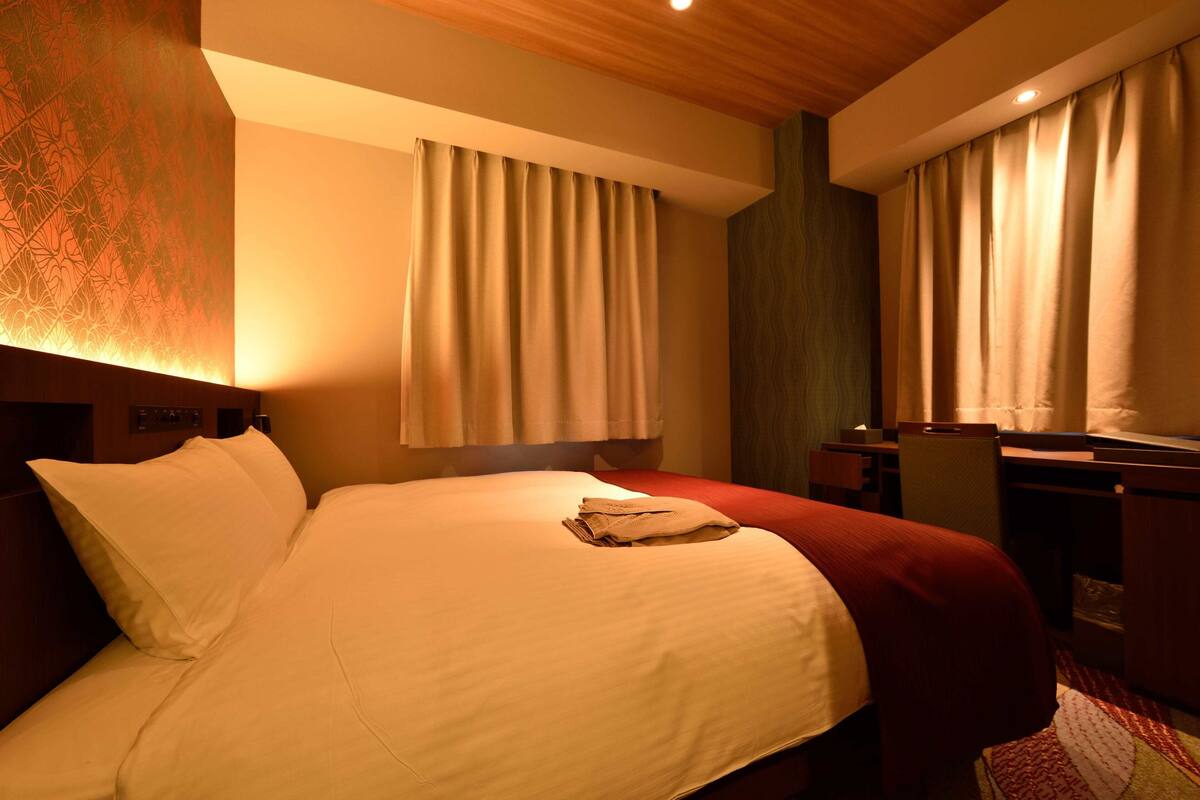 A hotel room with a large bed, two windows with curtains, a desk, and a chair.