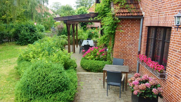 A brick building with a patio area featuring a table and chairs, surrounded by lush greenery and flowering plants. - Ferienwohnung Schuster (Garlstorf am Walde)