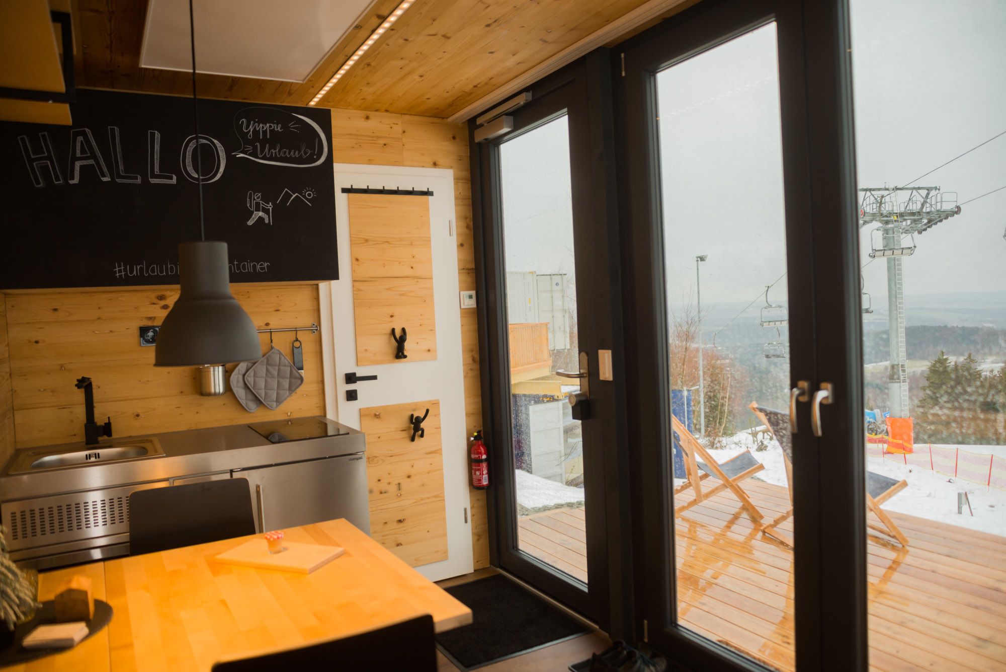 Cocina moderna con interior de madera, una pizarra con escritura y vistas a una terraza nevada a través de amplios ventanales.