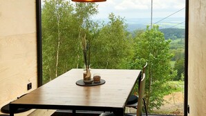 A dining area with a wooden table and chairs, a hanging pendant light, and a view of a forested landscape through large windows. - Bergheim Container Lodges (Schoeneck)