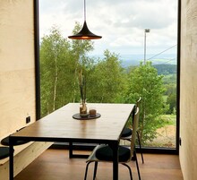 Un comedor con una mesa y sillas de madera, una lámpara colgante y vistas a un paisaje boscoso a través de amplios ventanales.