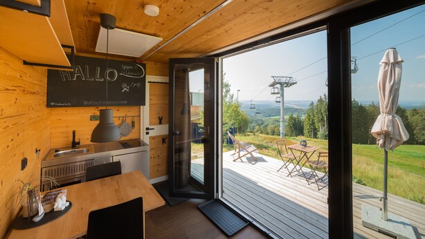 A modern kitchen with wooden walls, a large window, and a deck with outdoor furniture. - Bergheim Container Lodges (Schoeneck)
