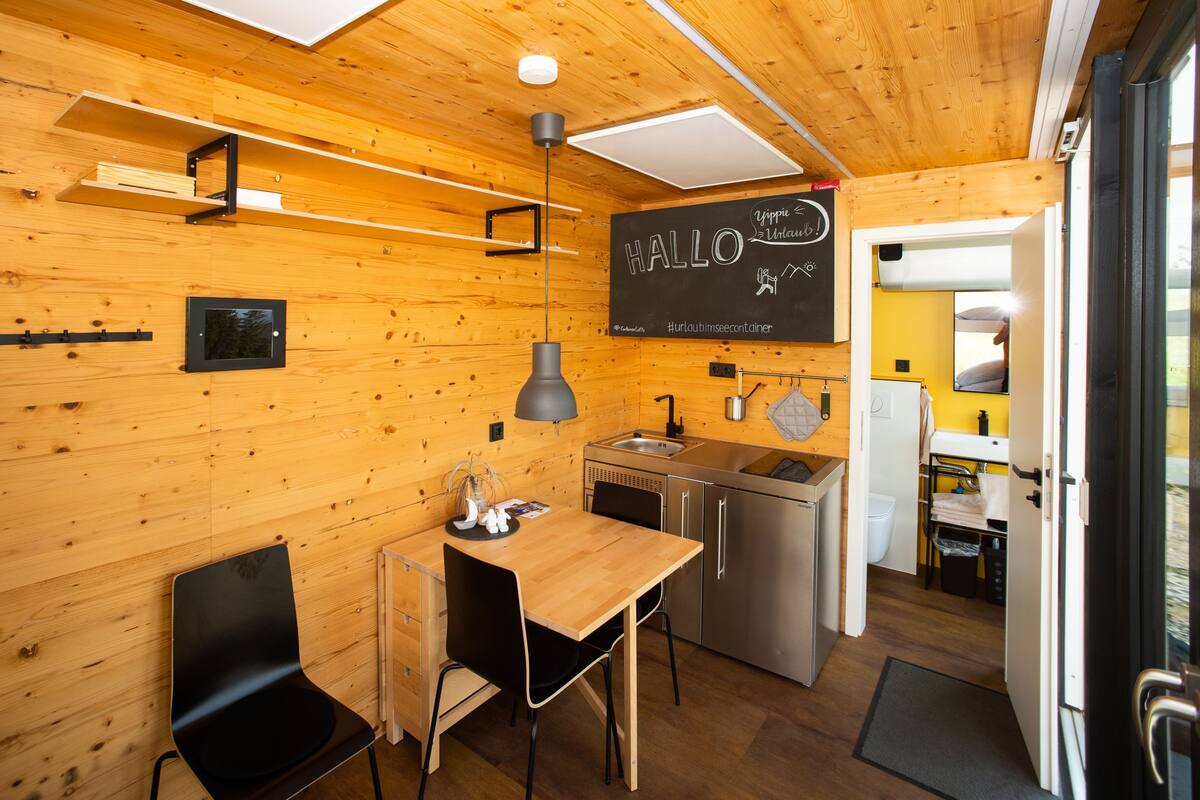 A compact kitchen with wooden walls, a blackboard with 'HALLO' and 'yippee! Wamb!' written on it, a sink, and a refrigerator.