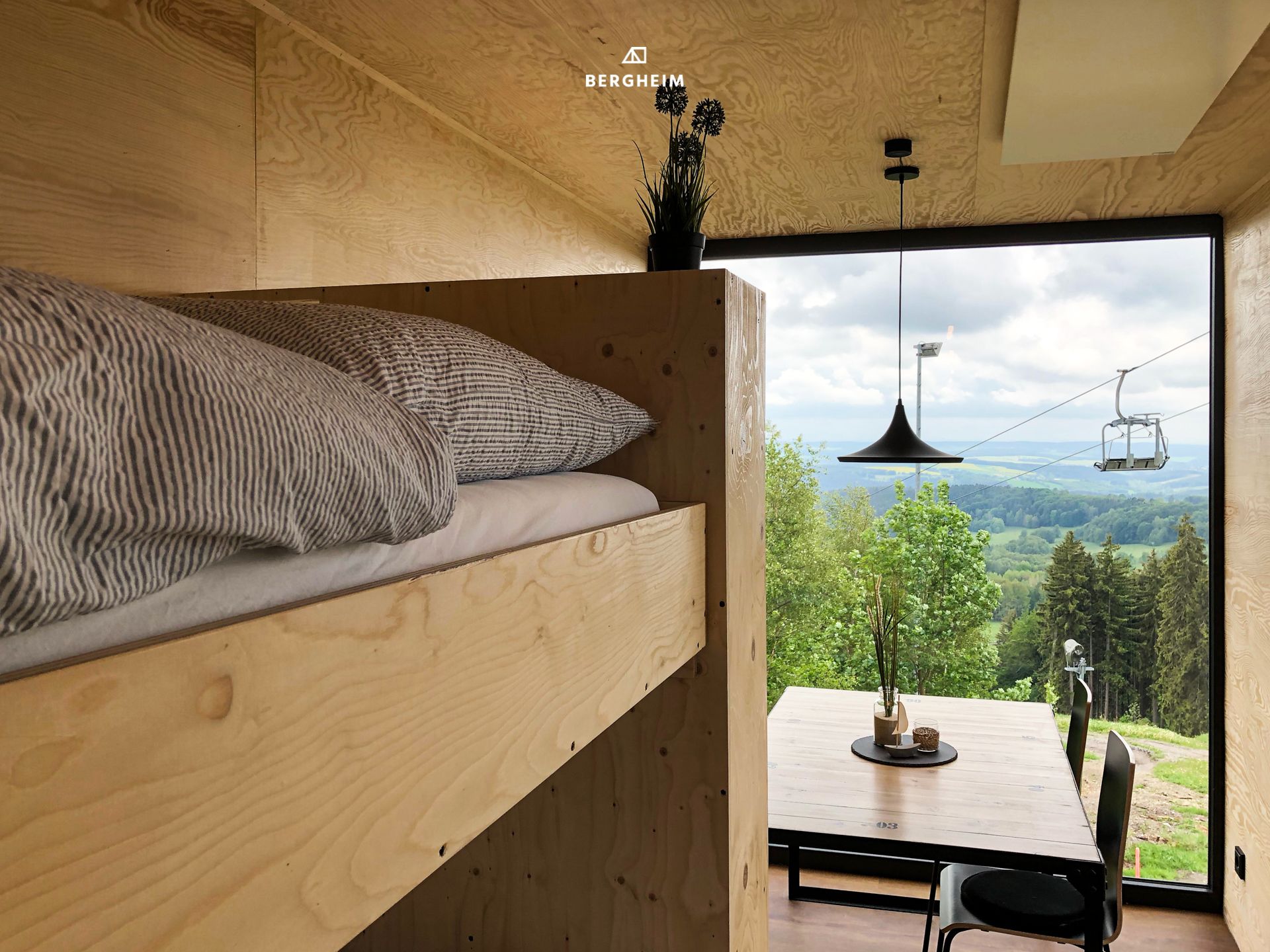 Una habitación moderna con una litera de madera, una zona de comedor con mesa y sillas, y un ventanal que ofrece vistas a un teleférico y al paisaje.