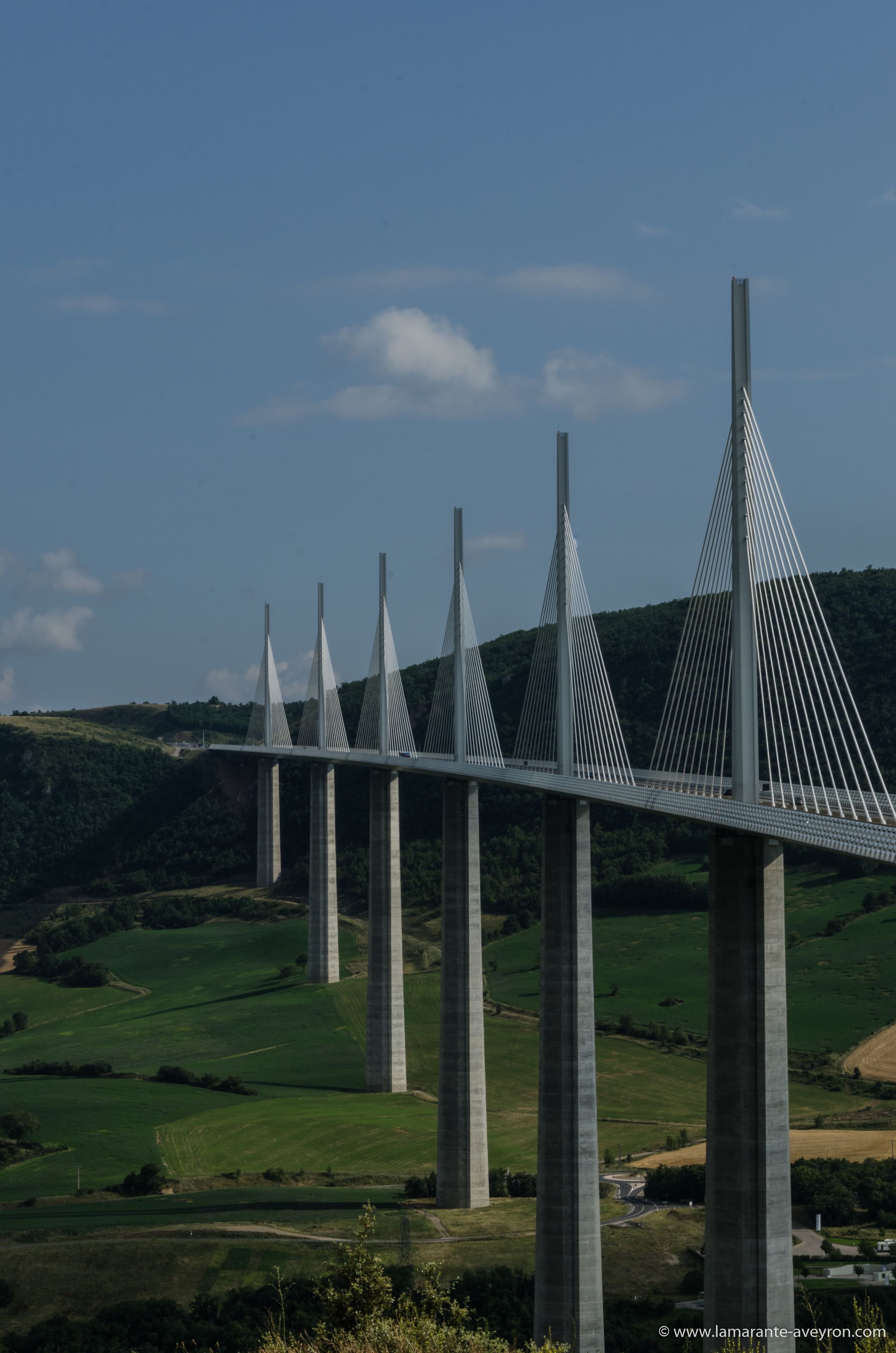 A large suspension bridge with multiple towers and cables, spanning over a green valley with hills in the background.