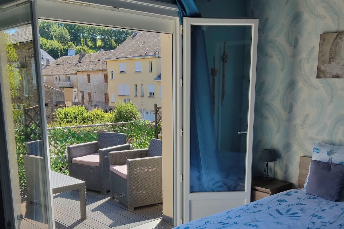 A bedroom with a bed, a balcony with wicker furniture, and a view of buildings and greenery.