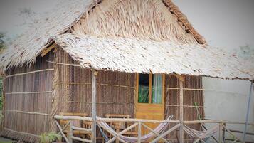 A rustic thatched-roof hut with a wooden porch, two hammocks, and a wooden door.