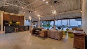 A spacious living area with a large open kitchen, a seating area with a view of the ocean, and a high ceiling with exposed beams. - Kutus Kutus Saba Beach Villa (Blahbatuh)