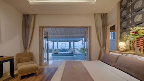 A bedroom with a large bed, a wooden table, a chair, and a view of an outdoor pool area. - Kutus Kutus Saba Beach Villa (Blahbatuh)