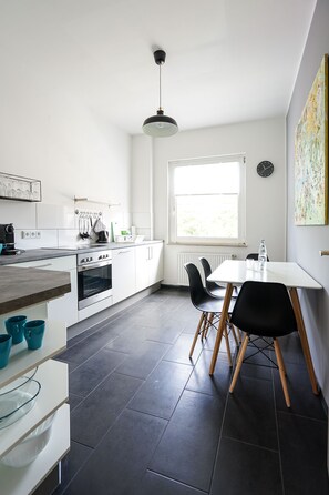 A modern kitchen with white cabinets, a stove, and a dining area with a table and chairs. - Glück Auf Appartements Rembrandtstraße (Essen)