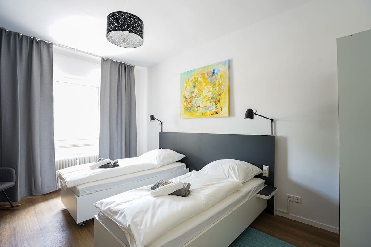 Ein modernes Hotelzimmer mit zwei Einzelbetten, einem großen Fenster mit Vorhängen, einem Gemälde an der Wand und einer Deckenleuchte.