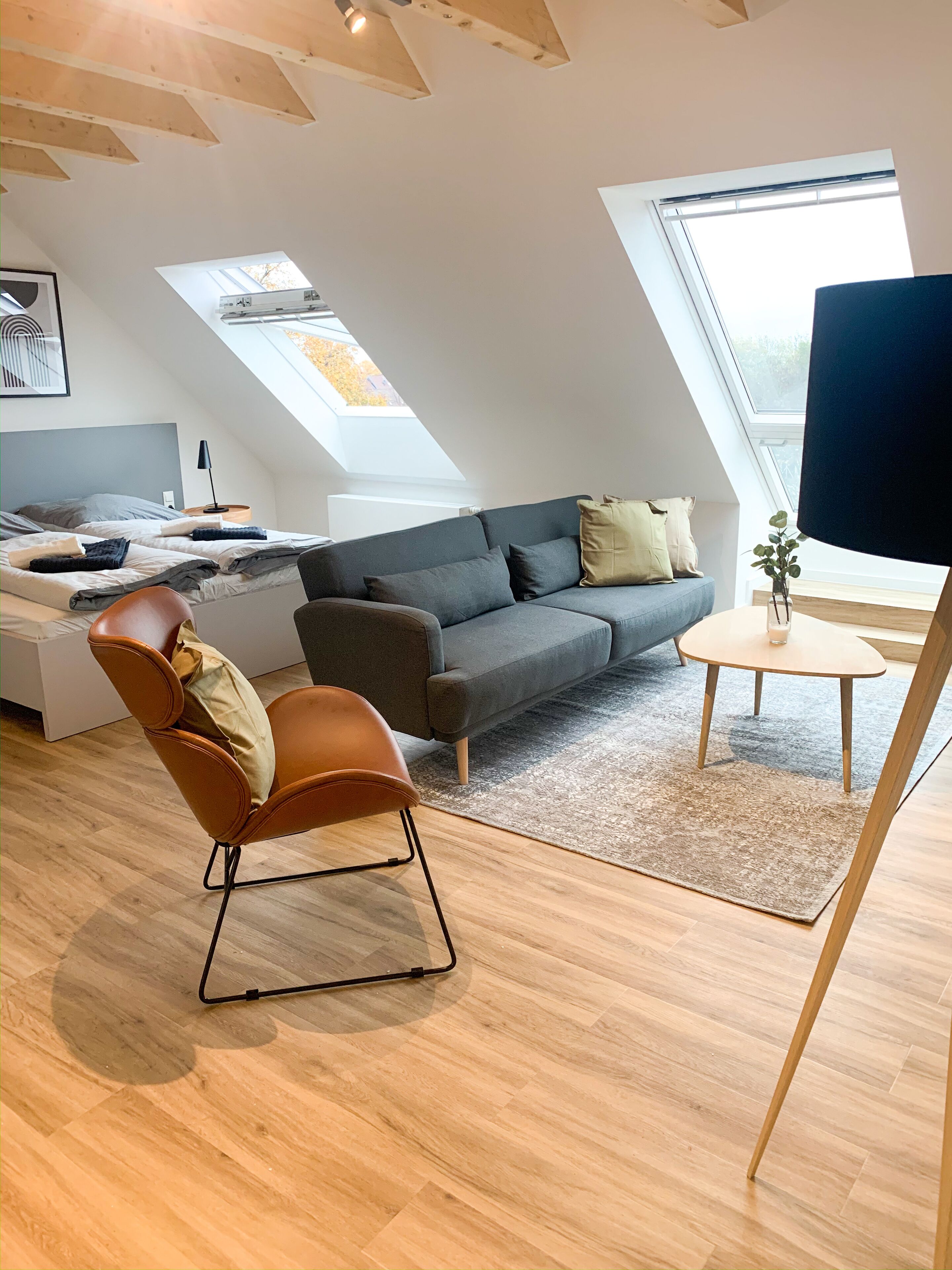 Ein modernes Loft-Zimmer mit schrägem Dach, einem grauen Sofa, einem braunen Ledersessel, einem kleinen runden Tisch und einem Oberlicht.