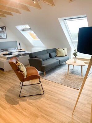 A modern loft-style room with a sloped ceiling, a gray sofa, a brown leather chair, a small round table, and a skylight. - Glück Auf Appartements Rembrandtstraße (Essen)