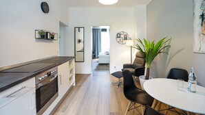 A modern kitchen with a built-in oven, a dining area with a round table and black chairs, and a living area with a sofa and a plant. - Glück Auf Appartements Rembrandtstraße (Essen)