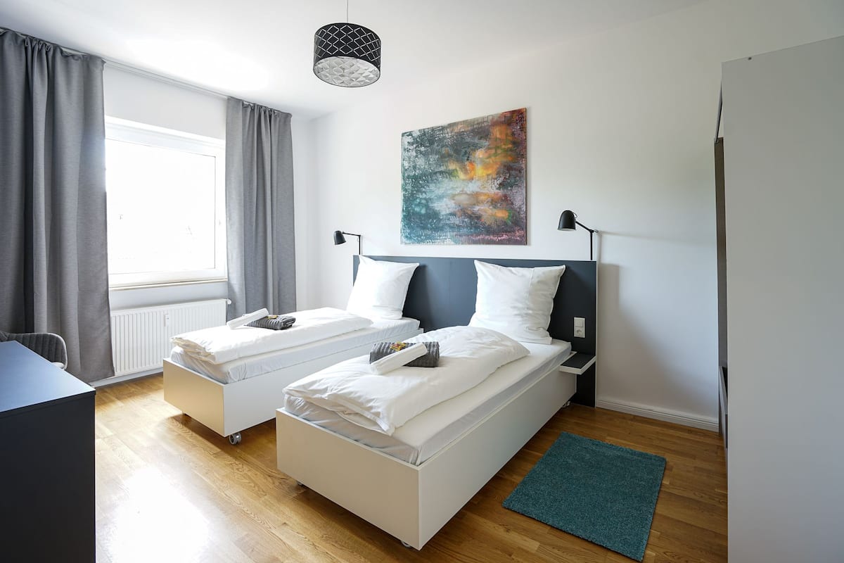 Ein Hotelzimmer mit zwei Betten, einem Schreibtisch, einem Fenster mit Vorhängen, einem Bild an der Wand und einer Lampe.