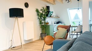 A modern living room with a grey sofa, wooden beams, a dining area with a table and chairs, a potted plant, and a kitchen in the background. - Glück Auf Appartements Rembrandtstraße (Essen)