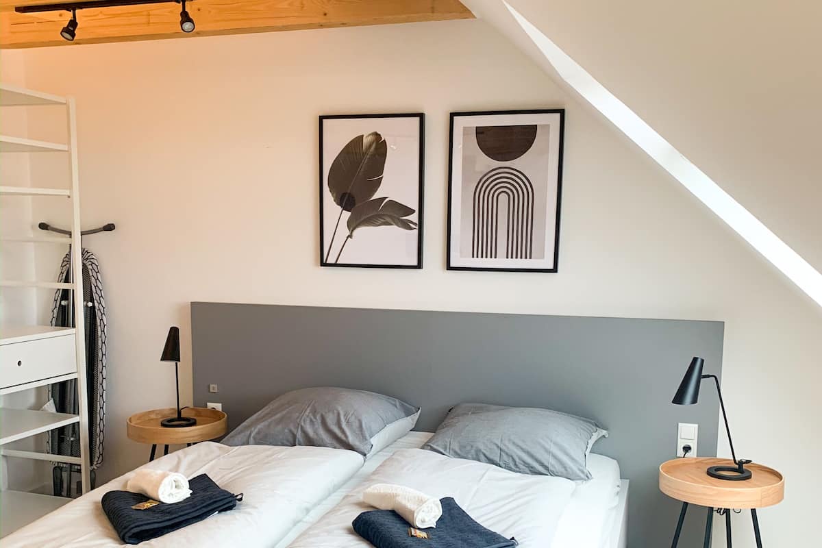 Ein modernes Schlafzimmer mit Schlafgalerie, einem Bett mit grauer Bettwäsche, zwei Kissen, einem Nachttisch mit Lampe und einem wandmontierten Regal.