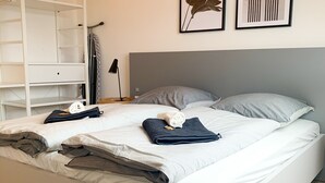 A modern bedroom with a white bed, grey pillows, and a grey headboard. There is a white wardrobe with open shelves and a black lamp beside the bed. - Glück Auf Appartements Rembrandtstraße (Essen)
