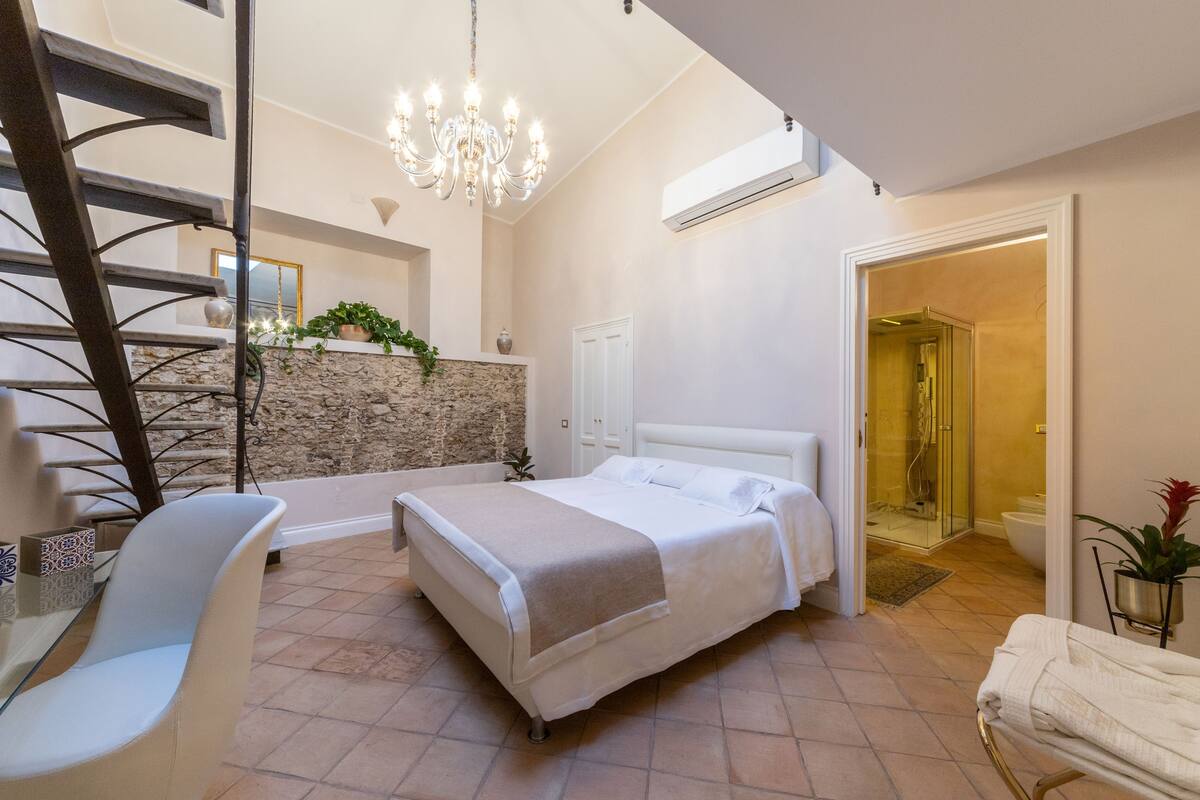 A bedroom with a bed, a chair, a staircase, a chandelier, and a bathroom with a shower.