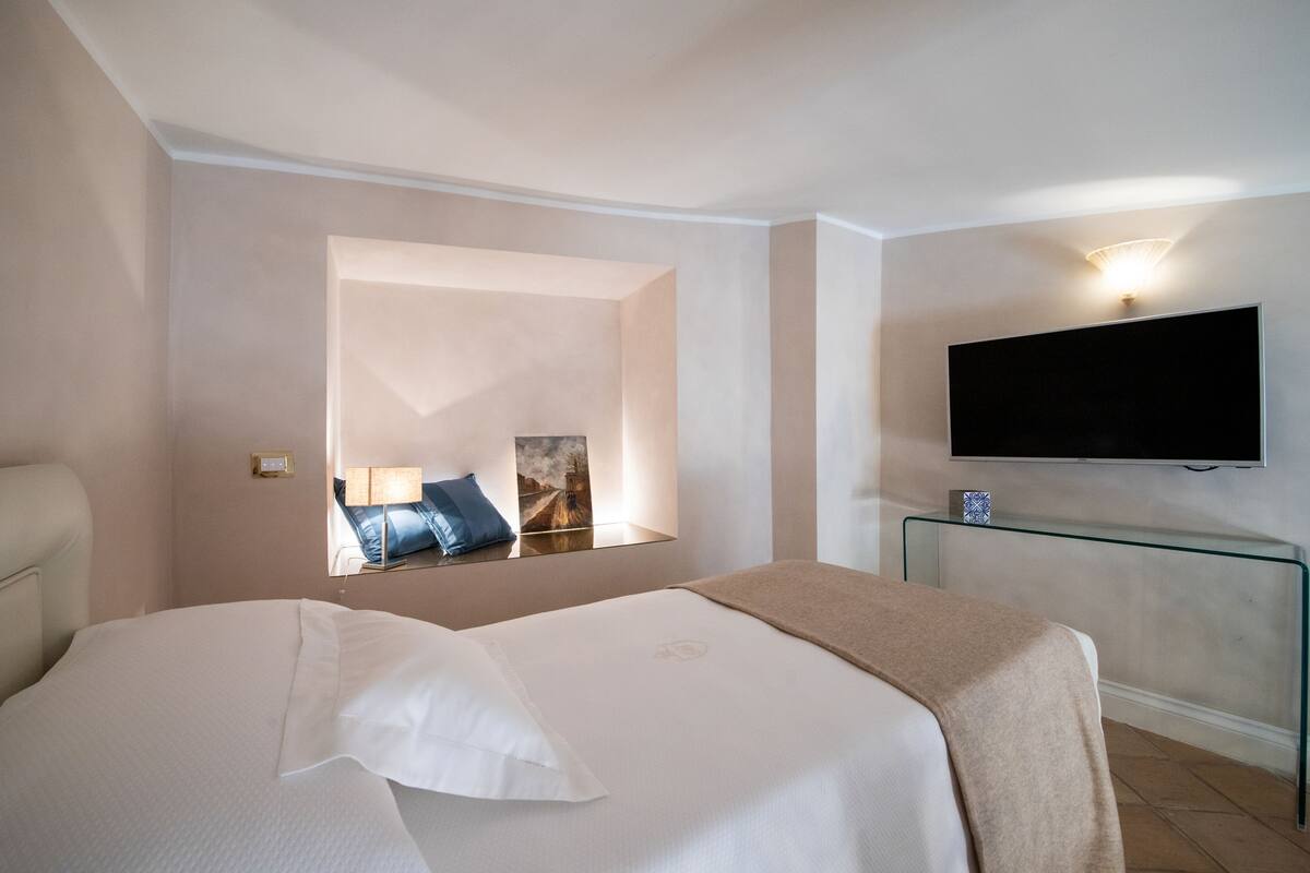 A hotel room with a bed, a TV mounted on the wall, a bedside table, and a mirror reflecting the room.