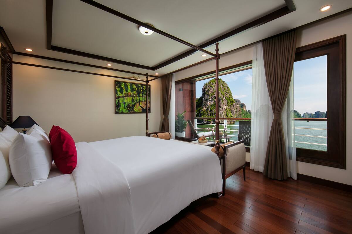 Quarto de hotel com uma cama grande, uma varanda com vista para um corpo de água e montanhas, e um quadro na parede.