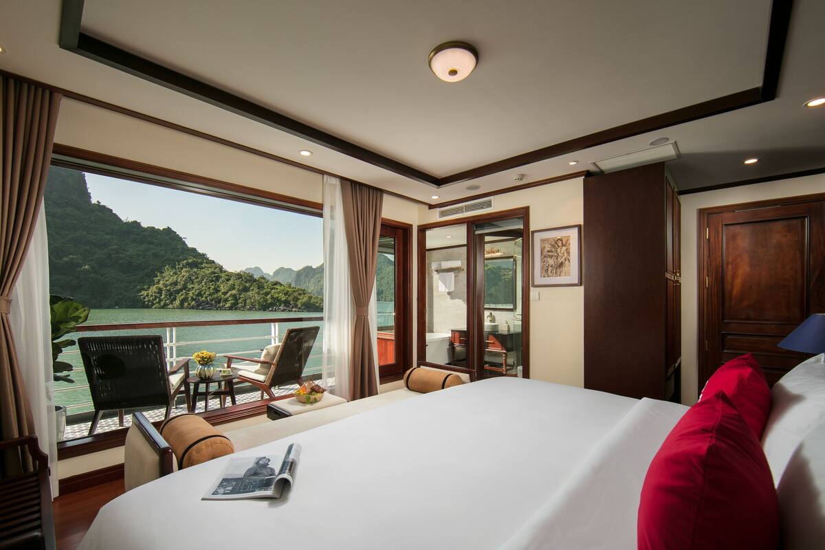 Quarto de hotel com uma cama grande, uma escrivaninha com cadeira, uma mesa pequena com um vaso de flores e vista para uma paisagem montanhosa através de janelas amplas.
