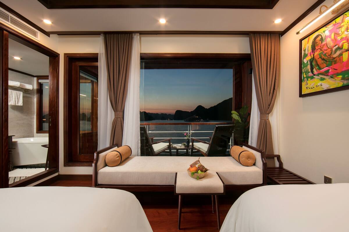 Quarto de hotel com duas camas, uma mesinha com uma cesta de frutas, vista para o mar e um quadro na parede.