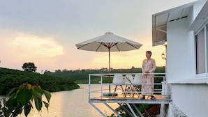 A balcony with a table and chairs, an umbrella, and a view of a river and greenery at sunset. - Ho Gia Trang (Tan Ha Lam Ha)