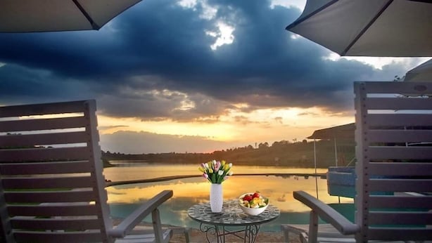 A table with a vase of flowers and a bowl of fruit on a balcony overlooking a lake with a sunset view. - Ho Gia Trang (Tan Ha Lam Ha)