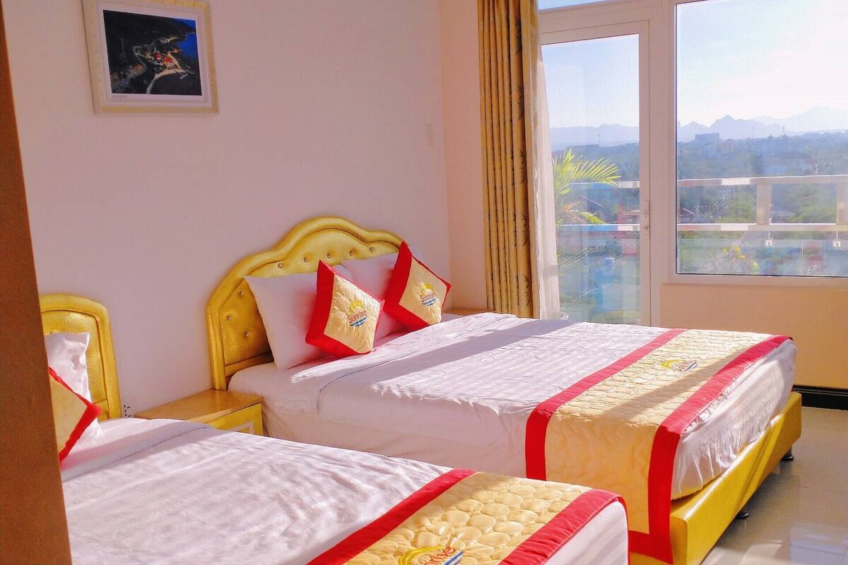 A hotel room with two beds, each with a yellow frame and white bedding. There is a window with a view of a cityscape and mountains.