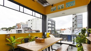 Un balcón con una mesa y sillas de madera, plantas en macetas y vistas de edificios de la ciudad.