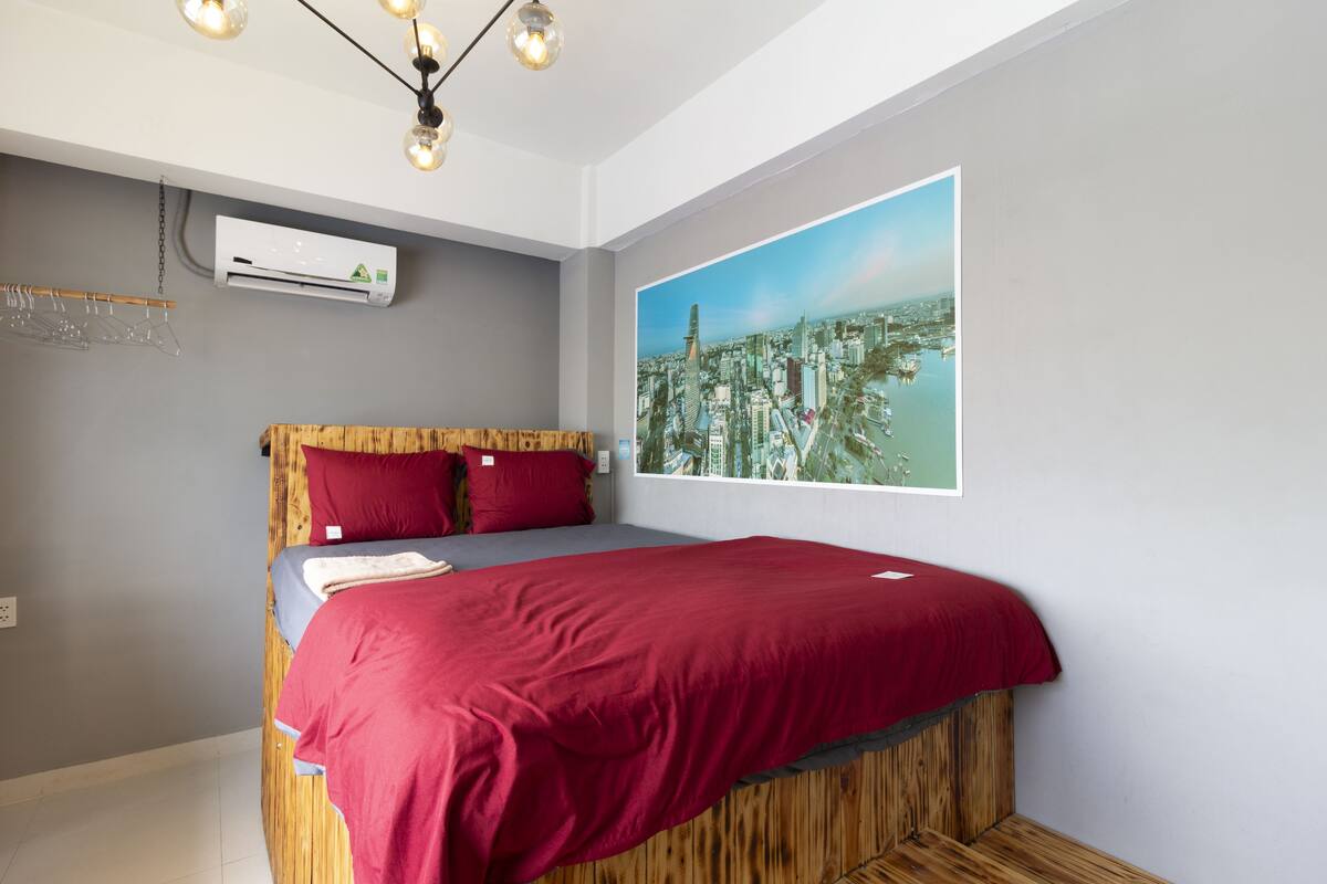 A bedroom with a bed, a cityscape view, an air conditioning unit, and a hanging light fixture.