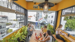 Un bar en la azotea con una mesa de madera, rodeado de plantas en macetas, con vistas a un paisaje urbano.