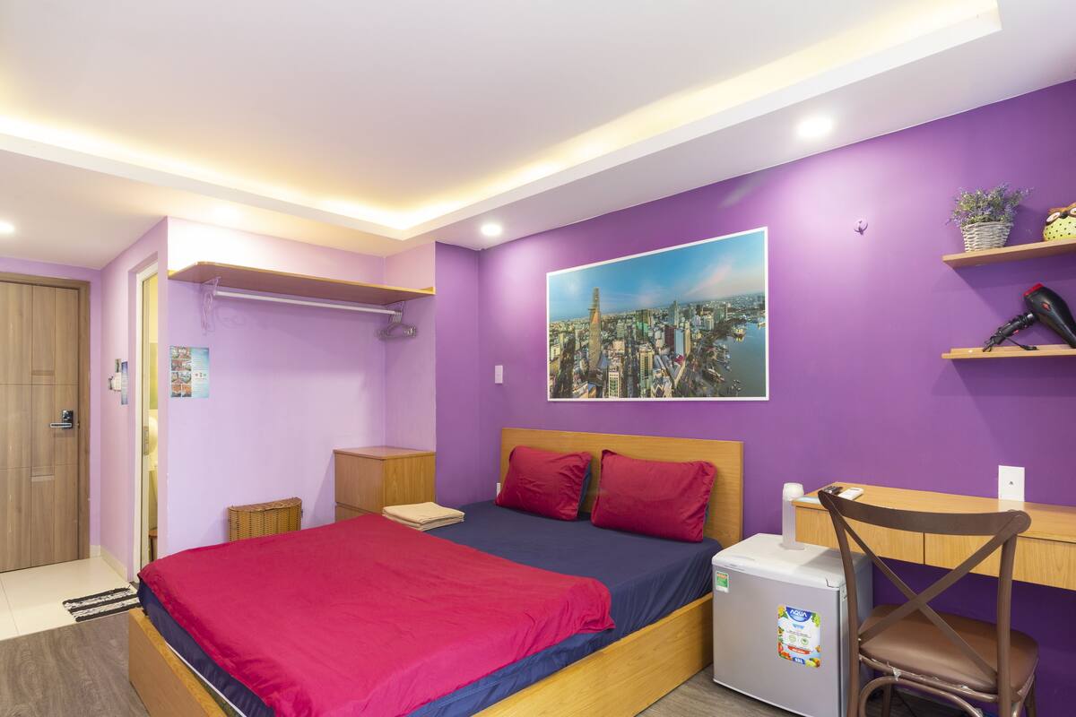 A bedroom with a bed, a desk, a chair, a refrigerator, and a cityscape painting on the wall.