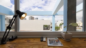 Una habitación con un ventanal grande, un escritorio con una lámpara, un control remoto, un periódico y una planta en maceta.