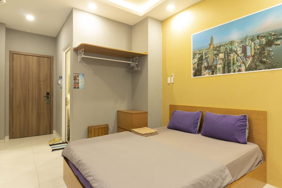 A bedroom with a bed, a wooden wardrobe, a small wooden nightstand, and a wall-mounted shelf.