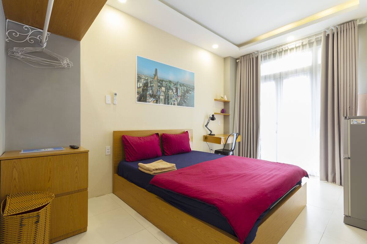 A modern hotel room with a large bed, a desk, a chair, a small table, a lamp, a refrigerator, and a cityscape painting on the wall.