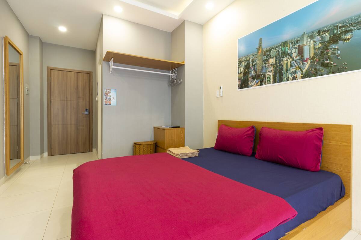 A bedroom with a bed, a wooden headboard, a cityscape painting, a wardrobe, and a small wooden cabinet.