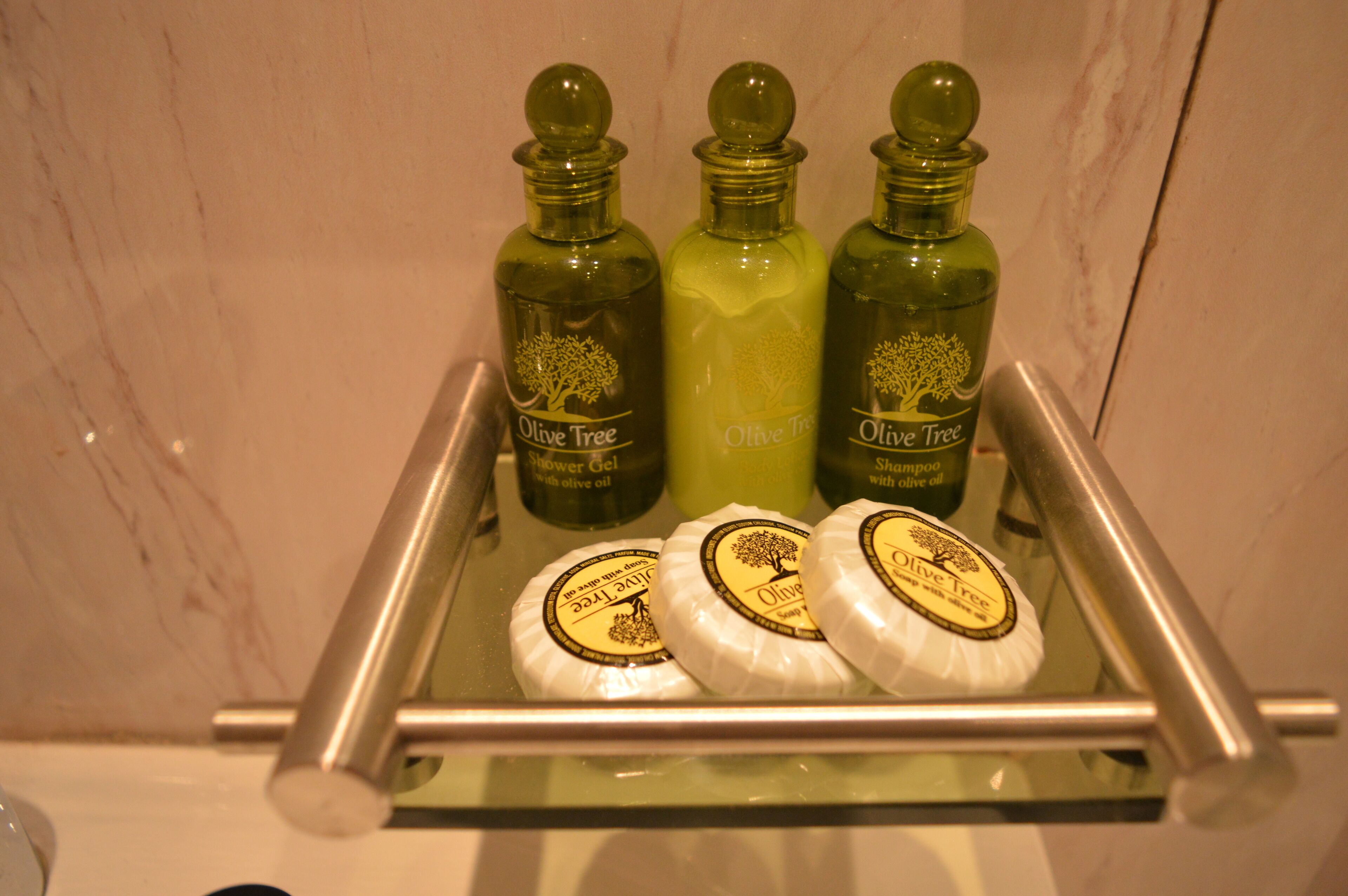 A bathroom shelf with three bottles of Olive Tree shower gel and two bars of soap.