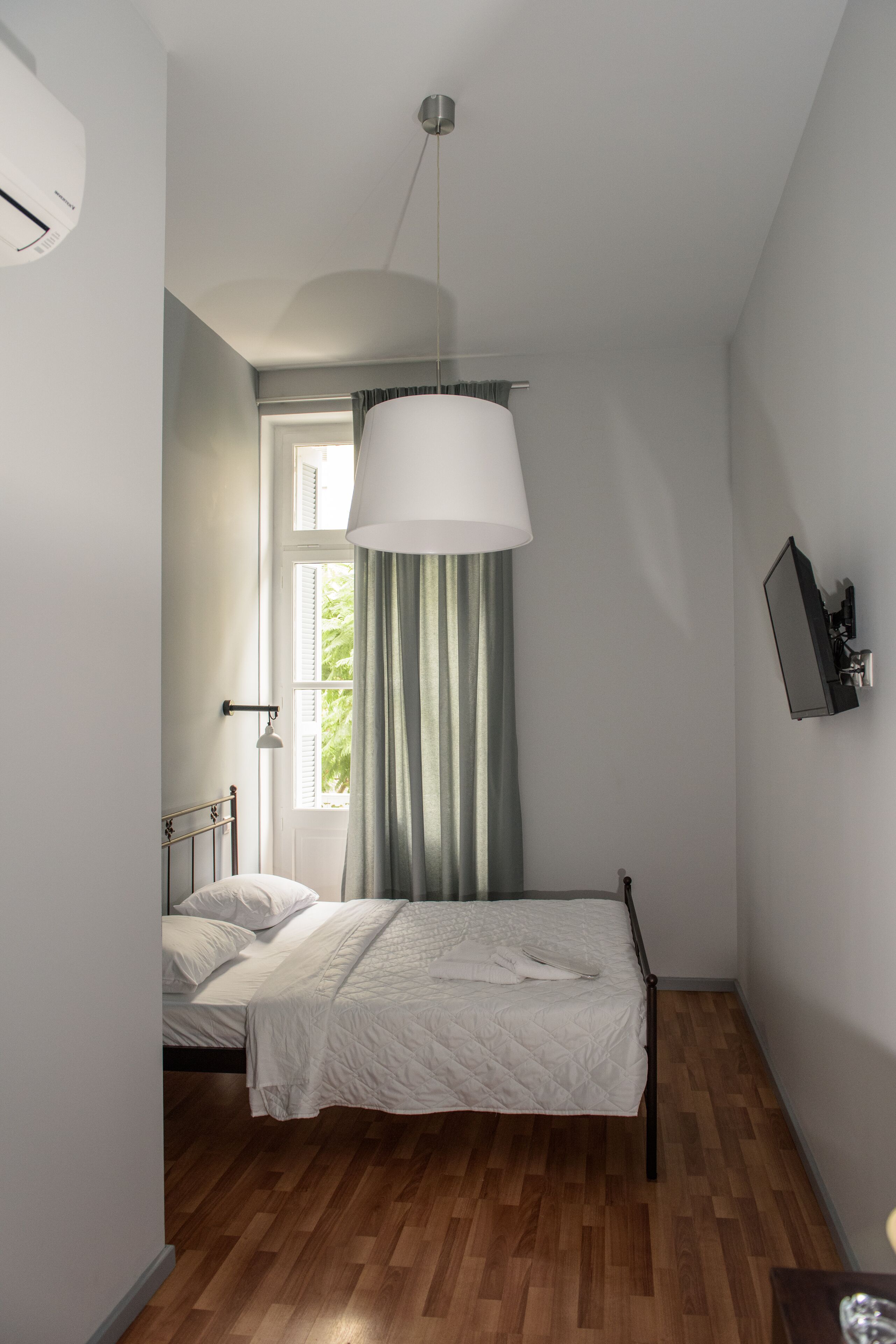 A bedroom with a bed, a window with curtains, a wall-mounted air conditioner, and a ceiling light fixture.