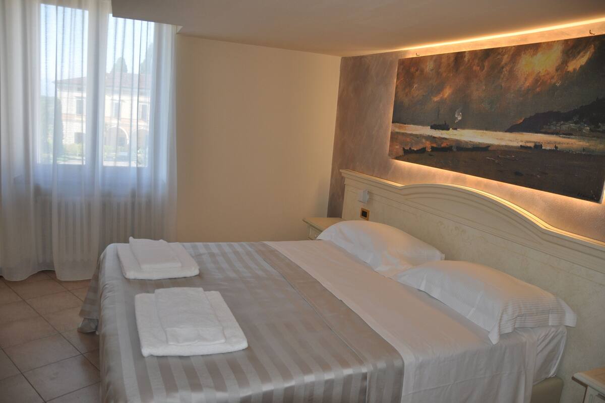 A hotel room with two beds, a painting on the wall, and a view of the sea through the window.